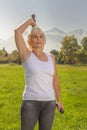 Elderly woman lifts dumbbells while doing fitness in a city park against the backdrop of mountains on a sunny day.