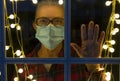 An elderly woman looks out the window sadly while wearing a medical mask due to the coronavirus. Self isolation and solitude away Royalty Free Stock Photo