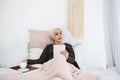 An elderly woman lying in bed drinking morning coffee uses a tablet to view news or chat with friends on a social Royalty Free Stock Photo