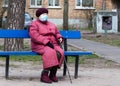 An elderly woman in a medical mask sits on a bench alone