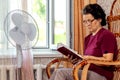 An elderly woman reads the Bible sitting in a chair by an open window and a fan Royalty Free Stock Photo