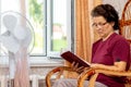 An elderly woman reads the Bible sitting in a chair by an open window and a fan Royalty Free Stock Photo