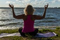 An elderly woman sitting in the lotus position and raising her hands up, on a mat in the park and looking at the lake Royalty Free Stock Photo