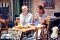 Elderly woman talking on her cell phone, smiling, while young adult female is looking at her with intriguing smile, drinking