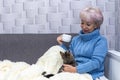 An elderly woman of 60-65 is warming up at the heating radiator with her cat, holding a white cup in her hand. Concept: low indoor Royalty Free Stock Photo
