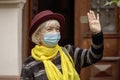 An elderly woman 60-65 years old in a medical mask greets someone by raising her hand . Concept: retirement, tourism and travel, f