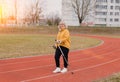 An elderly woman in a yellow sports jacket practices Nordic walking outdoors on the stadium`s rubber treadmill. A sunny sunset.