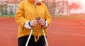 An elderly woman in a yellow sports jacket practices Nordic walking outdoors on the stadium`s rubber treadmill. A sunny sunset.