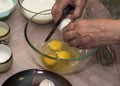 An elderly woman breaks an egg with a knife into a glass bowl for making pancake batter.Ingredients on the table-milk, salt, sugar Royalty Free Stock Photo