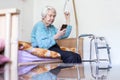 Elderly 96 years old woman reading phone message while sitting on medical bed supporting her by holder.