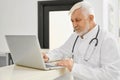 Eldery doctor in coat using laptop on reception.