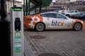 Electric car charging point with Tesla car plugged in decorated with Bitcoin and Nasdaq logotypes.