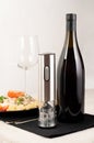 Electric corkscrew in gray metal. There is a bottle of wine, a plate of pasta, a glass and cutlery on the table next to it