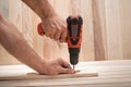 Electric drill screwdriver in male hand. Tightening screw, processing workpiece on light brown wooden table