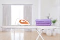 An electric iron stands on an ironing board and folded bath towels in Very Peri purple, against the backdrop of a home interior.