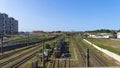 An electric locomotive stands on graveled and electrified railroad tracks. Behind a concrete fence are residential houses and a ci