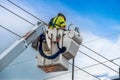 Electrician workman in green safety vest and helmet in crane lift working on highlines or traffic lights high in the air with sky