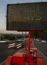 Electronic traffic sign stating Expect Delays with traffic blurred at rush hour