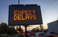 Electronic traffic sign stating Expect Delays with blurred traffic at sunset