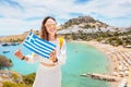 Asian woman in white dress posing with greek flag in front of the famous Lindos town on Rhodes island. Vacation and travel