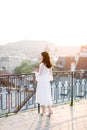 Elegant asian woman in white dress standing on rooftop terrace looking at city background skyline view of old European