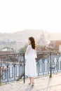 Elegant asian woman in white dress standing on rooftop terrace looking at city background skyline view of old European