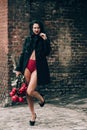 Elegant beautiful brunette woman in red shorts and black coat posing near brick wall with a big bouquet of red roses Royalty Free Stock Photo