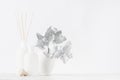 Elegant blank white glass bottles with aromatic sticks on soft light white wood table with silver decorative leaves as modern.