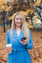 Elegant blonde mature woman texting on phone and drinking coffee in autumn park Royalty Free Stock Photo