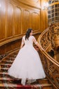 Elegant bride in white dress with long tail at old vintage palace walking up big wooden stairs