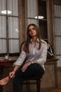 Elegant cute young woman in a white fashionable elegant shirt in stylish black jeans sits on a wooden chair in a vintage room. Royalty Free Stock Photo