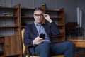 Elegant elderly man in glasses working on his mobile phone and sitting on chair in office Royalty Free Stock Photo