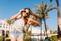 Elegant girl with beautiful hairstyle in sunglasses and denim shorts gladly posing in front of building and palm trees