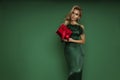 Elegant glamour blonde woman in evening feather green dress is holding red Christmas gift on green background in studio