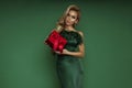 Elegant glamour blonde woman in evening feather green dress is holding red Christmas gift on green background in studio