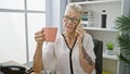 Elegant grey-haired senior woman boss engrossed in a serious phone conversation, relaxedly sipping coffee at her office Royalty Free Stock Photo