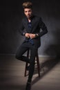 Elegant guy posing seated in dark studio fixing his jacket