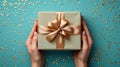 Elegant hands holding a gift box with a golden ribbon, presented against a turquoise backdrop sprinkled with golden confetti