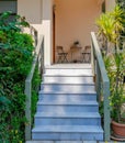An elegant house entrance marble stairs between foliage and a small terrace with table and chairs.