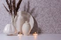 Elegant hygge home decor in evening with burning candles, white ceramic bottles, bowl, plate and bouquet of dried plant on white.