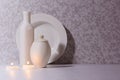 Elegant hygge home decor in evening with burning candles, white ceramic bottles, bowl, plate on white wood table in moonlight.