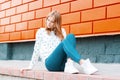 Elegant modern young cheerful woman a blonde in a white sweater in trendy jeans in sneakers sits on the ground near Royalty Free Stock Photo