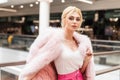 Elegant portrait pretty luxurious young woman with blond hair in chic pink expensive fur coat in stylish white top in mall. Sexy