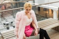 Elegant portrait pretty luxurious young woman with blond hair in chic pink expensive fur coat in stylish white top rests in mall.