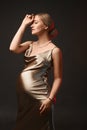 An elegant pregnant lady in a gold evening dress stands in front of a black background