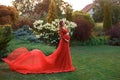 An elegant, pregnant woman walks in a beautiful garden in a luxurious, expensive red dress with a long train. Artistic