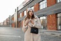 Elegant pretty stylish young woman in a vintage hat in a trendy coat with a fashion black leather handbag posing standing in a Royalty Free Stock Photo