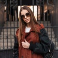 Elegant pretty young hipster woman in stylish sunglasses in fashionable brown trench coat with trendy leather black backpack walks Royalty Free Stock Photo