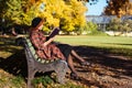 redhead woman in checked coat and black beret reading a book on bench, resting in autumn park at sunny day Royalty Free Stock Photo