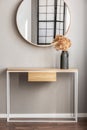 Elegant round mirror in wooden frame above fancy console table with flowers in vase in trendy bedroom interior with beige vase